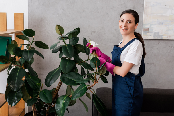 Side view of smiling cleaner holding spray bottle near plant at home  - Φωτογραφία, εικόνα