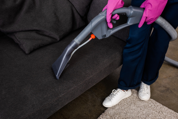 Cropped view of cleaner in rubber gloves holding vacuum cleaner near couch  - 写真・画像