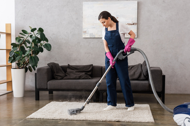 Hermosa limpiadora en ropa de trabajo y guantes de goma usando aspiradora con vapor caliente en la alfombra en la sala de estar
 - Foto, Imagen
