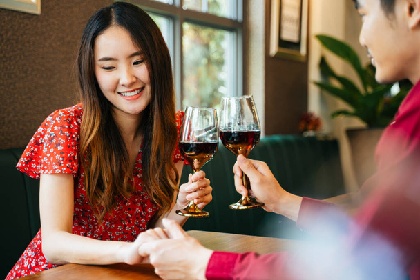 Vieren op Valentijnsdag. Mooie Aziatische vrouw in rode jurk toasten rode wijnglazen met haar vriendje. Hand op tafel houden. - Foto, afbeelding
