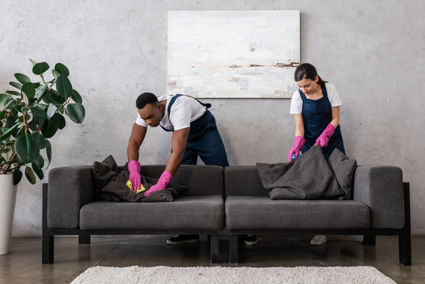 Мультикультурные чистящие средства в униформе диван для уборки с тряпками в гостиной
 - Фото, изображение