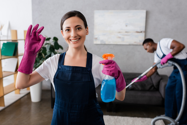 Focus selettivo di pulizia sorridente mostrando gesto ok e tenendo detergente vicino collega afro-americano con aspirapolvere a casa  - Foto, immagini
