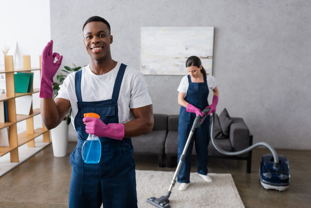 Selective focus of smiling african american cleaner showing okay and holding detergent near colleague using vacuum cleaner  - Photo, Image