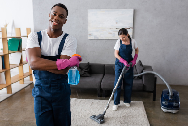 Concentration sélective de nettoyant afro-américain tenant le détergent et souriant à la caméra près de collègue nettoyage tapis  - Photo, image