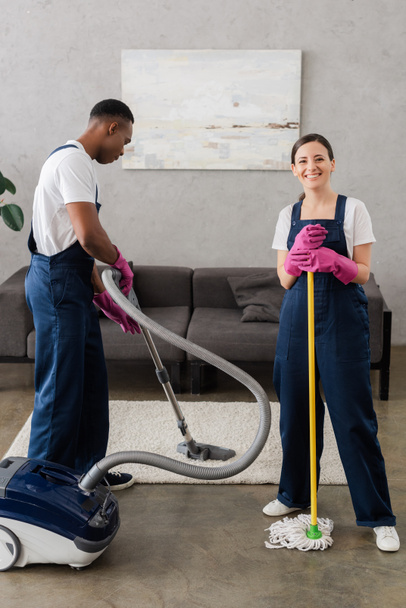 Limpiador sonriente sosteniendo fregona cerca de un colega afroamericano limpiando alfombras en casa
  - Foto, imagen