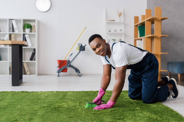 Smiling african american cleaner holding brush while cleaning carpet in office  - Photo, Image