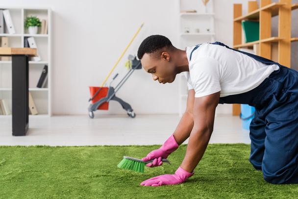 Vue latérale de nettoyant afro-américain en uniforme et gants en caoutchouc nettoyage tapis avec brosse dans le bureau  - Photo, image
