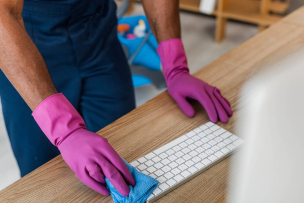 Ofisteki bilgisayar klavyesini temizleyen lastik eldivenli Afro-Amerikan temizlikçi görüntüsü.  - Fotoğraf, Görsel