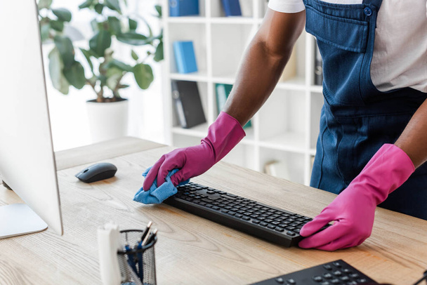 Cropped view of african american cleaner in rubber gloves cleaning computer keyboard on office table  - Photo, Image