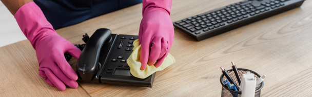 Panoramische oogst van Afrikaanse Amerikaanse reiniger in rubber handschoenen schoonmaken telefoon met doek in het kantoor  - Foto, afbeelding