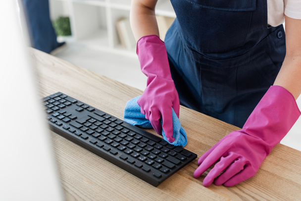 Widok czyszczenia w gumowych rękawiczkach trzymając szmatę w pobliżu klawiatury komputera w biurze  - Zdjęcie, obraz