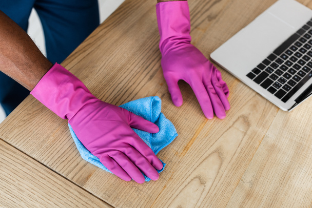 Widok przycięty afrykański amerykański środek czyszczący w gumowych rękawiczkach stół czyszczący w pobliżu laptopa w biurze  - Zdjęcie, obraz
