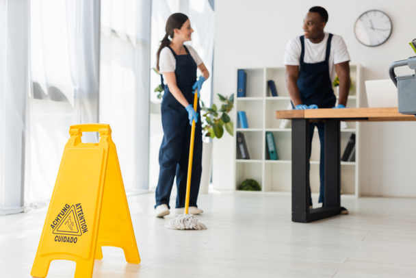 Selective focus of wet floor sign and smiling multiethnic cleaners working in office  - 写真・画像