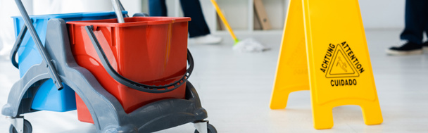 Cultura panorâmica do carrinho de limpeza perto do sinal do chão molhado e limpadores que trabalham no escritório
  - Foto, Imagem