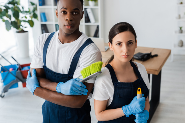 Multiethnic cleaners holding brush and mop while looking at camera in office  - Foto, imagen