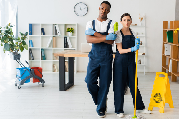Multicultural cleaners with mop and brush looking at camera near wet floor sign in office  - Foto, Imagem