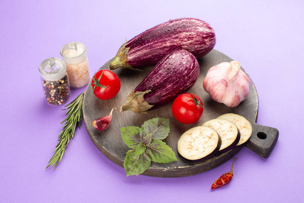 Summer vegetables on wooden board on purple background:ripe eggplants,tomatoes,fresh rosemary,purple basil leaves,garlic and hot pepper.Close-up. - Foto, imagen