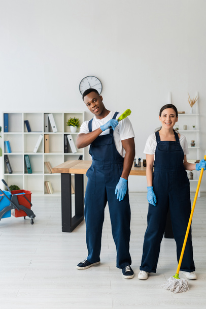 Positive multiethnic cleaners with brush and mop looking at camera in office  - Zdjęcie, obraz