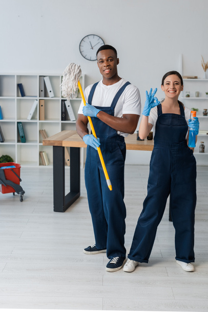 Smiling multiethnic cleaners holding mop and detergent and showing ok gesture in office  - Фото, зображення