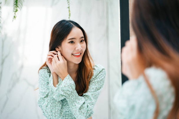 Schöne asiatische Thailänderin auf grünem Modehemd mit Ohrringen vor dem Spiegel. - Foto, Bild
