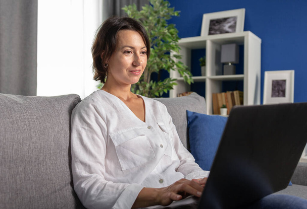Smiling woman relaxing on grey couch with wireless laptop - Фото, изображение