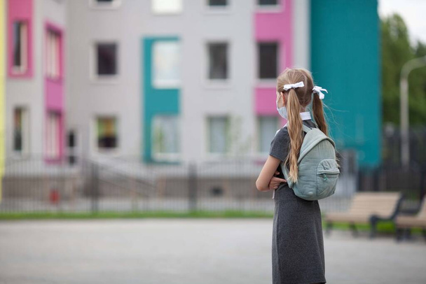 Девочка-подросток с рюкзаком остается рядом со школой. Вид сзади. Школьница в медицинской маске смотрит на своего школьного приятеля. Дети возвращаются в школу после пандемии
.  - Фото, изображение
