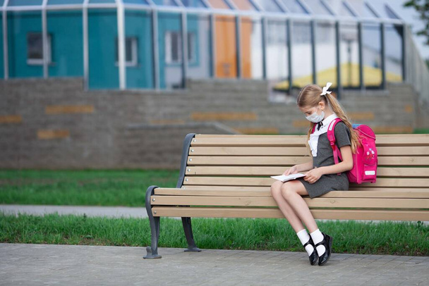 Une adolescente assise sur un banc ferme une école en regardant dans son cahier, répète ses devoirs. Une écolière portant un masque médical. Les enfants retournent à l'école après une pandémie. - Photo, image