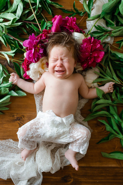 Una niña de dos meses yace sobre una mesa de madera con peonías y gritos. - Foto, Imagen
