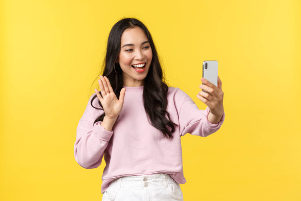 İnsanların duyguları, yaşam tarzı, eğlence ve güzellik konsepti. Arkadaşlarıyla görüntülü konuşma yapan sevimli Asyalı kız, el sallayarak telefon kamerasına merhaba diyor, blogcu canlı yayında, sarı arka planda. - Fotoğraf, Görsel