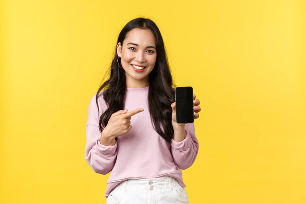 Pessoas emoções, lazer estilo de vida e conceito de beleza. Sorrindo mulher asiática 20s, mostrando exibição de smartphone, recomendo aplicativo ou jogo móvel, apontando o dedo para a tela, fundo amarelo - Foto, Imagem