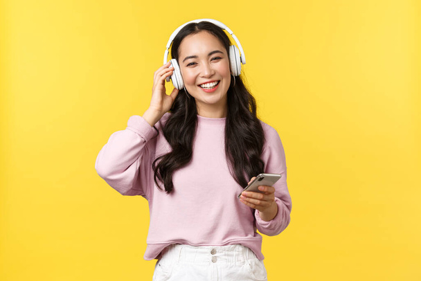 Mensen emoties, lifestyle vrijetijdsbesteding en schoonheid concept. Gelukkig glimlachende aziatische vrouw luisteren muziek in draadloze hoofdtelefoon en kijken blij met mooi geluid, met een mobiele telefoon - Foto, afbeelding