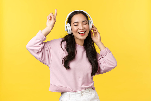 People emotions, lifestyle leisure and beauty concept. Pretty happy asian woman in wireless headphones, listening music and dancing, singing along, enjoying summer, stand yellow background - Фото, изображение