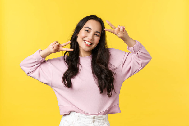 People emotions, lifestyle leisure and beauty concept. Cheerful and optimistic asian girl in stylish clothes, showing peace signs and smiling broadly, enjoying summer, yellow background - Photo, Image