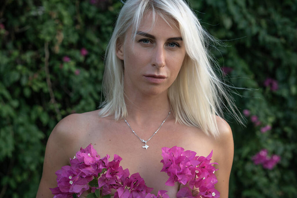 Portrait of young blonde woman with pink flowers cover breast and green bush with pink flowers on background - Φωτογραφία, εικόνα