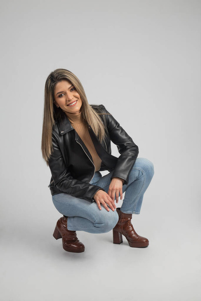 латиноамериканская блондинка в джинсах и черной кожаной куртке с красивыми сапогами, улыбающаяся  - Фото, изображение