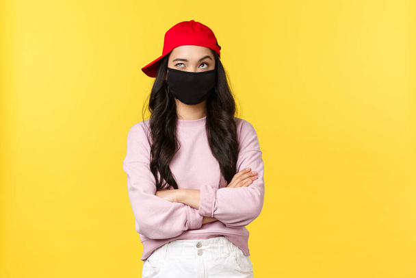 Covid-19, social-distancing lifestyle, prevent virus spread concept. Thoughtful and creative cute asian girl in red cap and face mask, looking up thinking and smiling with eyes, cross arms chest - Фото, изображение