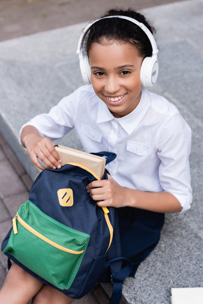 happy african american schoolgirl in headphones putting book in backpack - Foto, Imagem