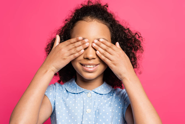 smiling cute curly african american kid covering eyes isolated on pink - Foto, Imagen