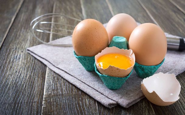 Свежие сырые куриные яйца в коробке с яйцами. Разбитое яйцо, желток. Органическая пища для хорошего здоровья с высоким содержанием белка на деревенском деревянном фоне
. - Фото, изображение