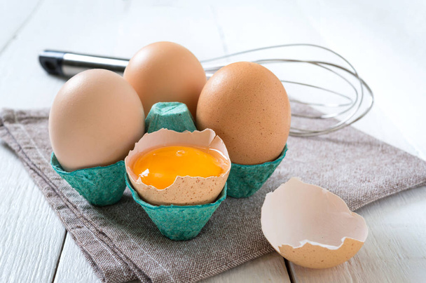 Świeże surowe jaja kurze w pudełku po jajkach. Rozbite jajko, żółtko. Żywność ekologiczna dla dobrego zdrowia wysokie białko na białym drewnianym tle. - Zdjęcie, obraz
