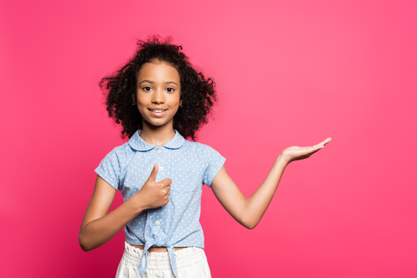 smiling cute curly african american kid pointing with hand aside and showing thumb up isolated on pink - Фото, изображение