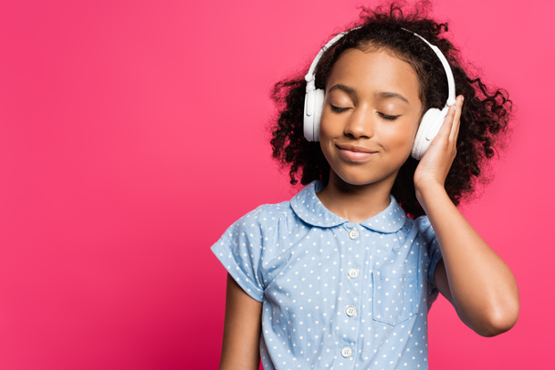 smiling curly african american kid in headphones with closed eyes isolated on pink - Fotografie, Obrázek