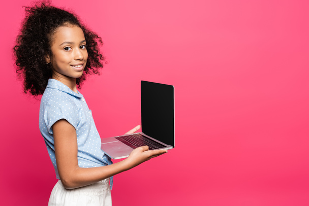 usmívající se roztomilé kudrnaté africké americké dítě drží notebook s prázdnou obrazovkou izolované na růžové - Fotografie, Obrázek