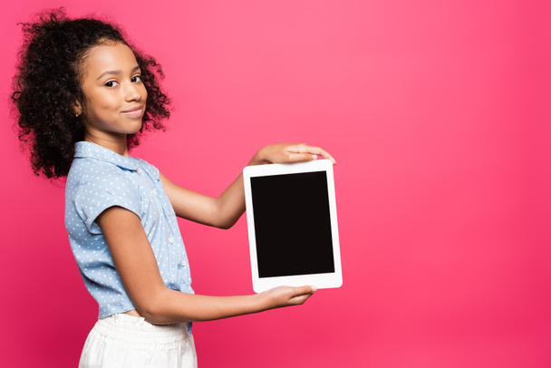 smiling cute curly african american kid presenting digital tablet isolated on pink - Фото, изображение