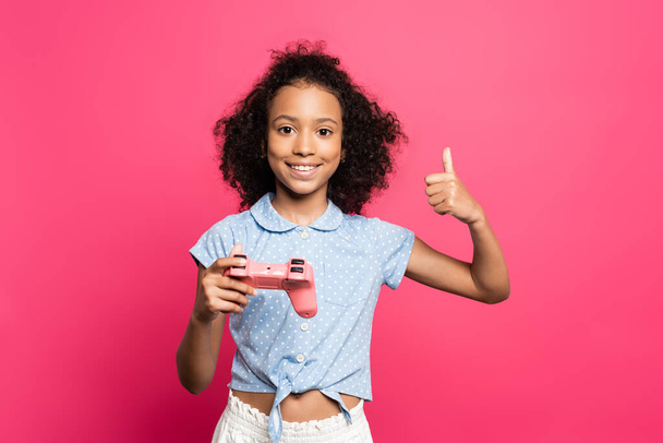 KYIV, UKRAINE - JUNE 9, 2020: smiling cute curly african american kid with joystick showing thumb up isolated on pink - Foto, Bild