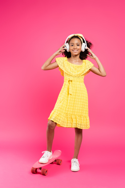 full length view of smiling curly african american child in yellow outfit and headphones on penny board on pink background - Fotografie, Obrázek