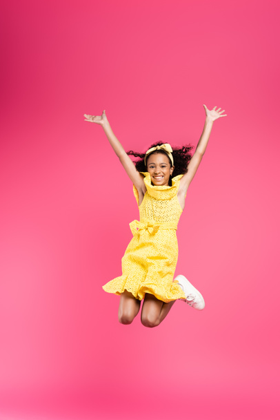 vue pleine longueur de heureux frisé afro-américain enfant en tenue jaune sautant avec les mains dans l'air sur fond rose - Photo, image