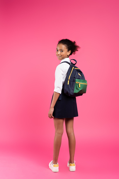 На задньому плані - усміхнена афроамериканська школярка з рюкзаком на рожевому фоні. - Фото, зображення