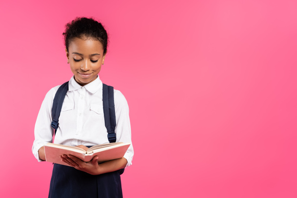smiling african american schoolgirl reading book isolated on pink - Foto, Imagem