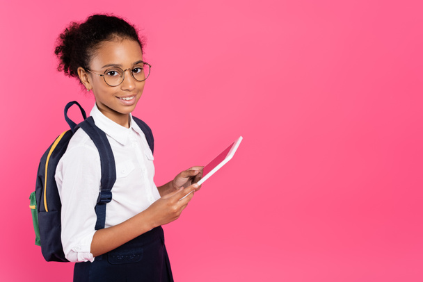 smiling african american schoolgirl in glasses with backpack using digital tablet isolated on pink - Zdjęcie, obraz
